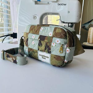 May include: A quilted cosmetic bag with a camouflage pattern, daisy accents, and a gold zip. The bag has a matching strap and the text "ArtsieFartsie Haus". A Brother sewing machine is in the background.