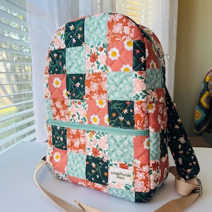 May include: A quilted patchwork backpack with a floral pattern in shades of coral, teal, and dark green. The backpack has a zippered front pocket, adjustable straps, and the text "ArtsieFartsie Haus".