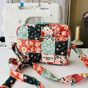 May include: A quilted crossbody bag with a patchwork design featuring floral patterns in red, white, and teal. The bag has a teal zipper and a long strap. The label reads "ArtsieFartsie Haus". A Brother sewing machine is in the background.
