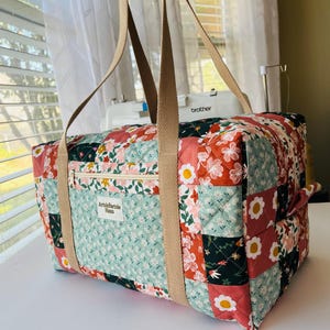 Puede incluir: Un bolso tote acolchado con un diseño de patchwork con estampados florales en rojo, rosa, verde y blanco. El bolso tiene una cremallera beige, asas beige y el texto "ArtisPartie Haus".