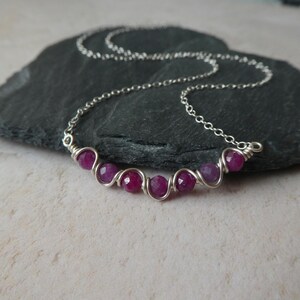 May include: A silver chain necklace featuring a horizontal bar design composed of five faceted, deep pinkish-red gemstone beads. Each bead is individually wrapped in thin silver wire, creating a continuous, interconnected link structure.