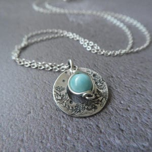 May include: A silver necklace with a pendant. The pendant has a light blue gemstone held by silver wire, set within a circular design with floral and dot patterns. The necklace chain is silver. The pendant's diameter is approximately 2.5 cm.