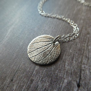 May include: A silver necklace with a round pendant featuring a detailed leaf design. The pendant is textured and has a slightly raised edge.