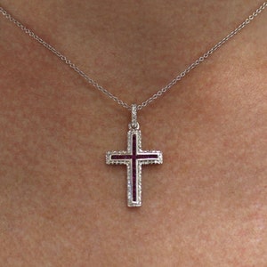 Pode incluir: Um colar de corrente de prata com um pequeno pingente de cruz de prata. A cruz tem um centro de esmalte roxo escuro e é cercada por pequenas pedras transparentes.