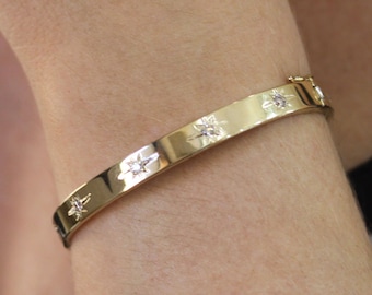 以下が含まれることがあります： 星型の小さなアクセントが並んだ金のバングルブレスレット。ブレスレットは人の手首につけています。