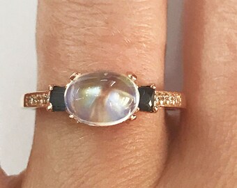 Puede incluir: Anillo de oro rosa con una piedra preciosa ovalada e iridiscente, flanqueada por dos piedras negras rectangulares. La banda está detallada con pequeños acentos brillantes, creando un diseño elegante.
