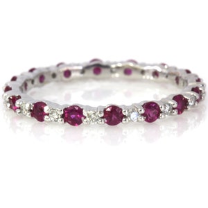 May include: A silver ring with a continuous band of round, deep red gemstones and small, clear accent stones. The ring is set against a white background, highlighting the sparkle of the gems. The ring is a piece of jewelry.