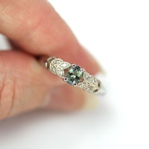 May include: A silver ring with a green gemstone surrounded by smaller white gemstones. The ring has a delicate leaf design.