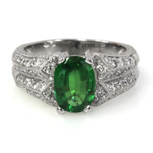 May include: A silver ring featuring a large oval green gemstone surrounded by smaller diamonds. The ring has an intricate design with a double band.
