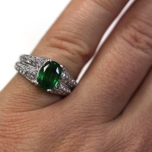 May include: A silver ring featuring a large oval emerald gemstone, surrounded by smaller diamonds. The ring is worn on a finger.