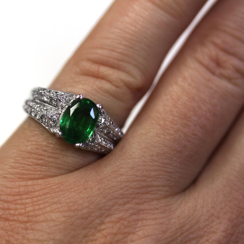 May include: A silver ring featuring a large oval emerald gemstone, surrounded by smaller diamonds. The ring is worn on a finger.