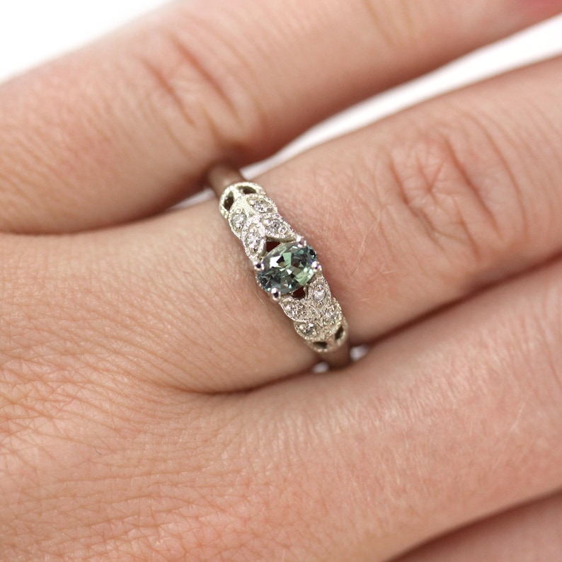 May include: A silver ring with a green oval gemstone surrounded by smaller white gemstones. The ring has a delicate, intricate design.