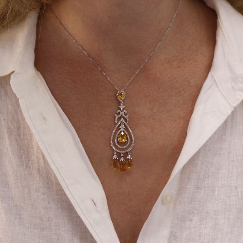 May include: A silver chain necklace with a dangling pear-shaped yellow gemstone pendant. The pendant is surrounded by smaller white gemstones and has a delicate, ornate design.