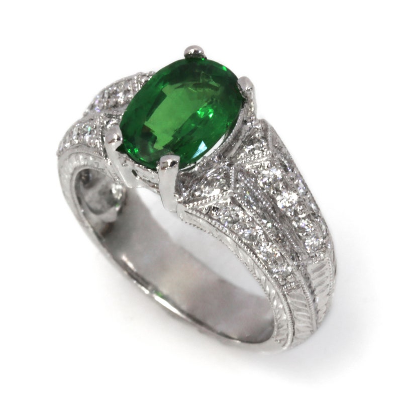 May include: A silver ring featuring a large oval green gemstone surrounded by smaller diamonds. The ring has an intricate design with a filigree pattern.