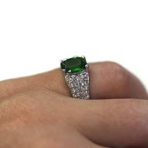 May include: A silver ring featuring a large, emerald green gemstone set in the centre. The ring is decorated with smaller, white gemstones around the band.