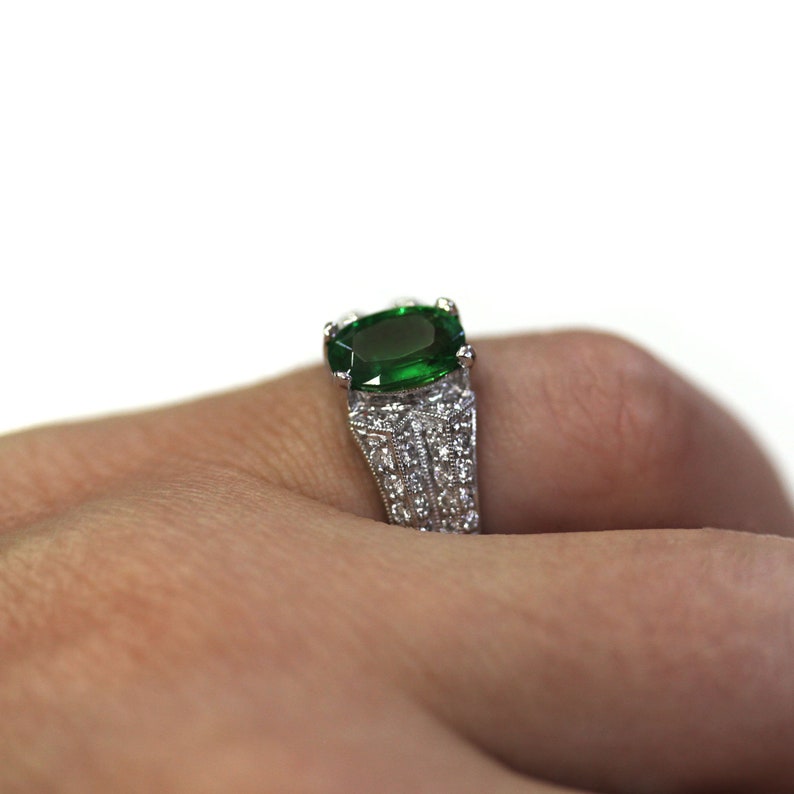 May include: A silver ring featuring a large, emerald green gemstone set in the centre. The ring is decorated with smaller, white gemstones around the band.