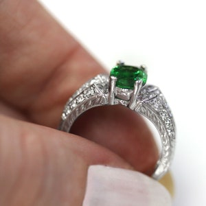 May include: A silver ring featuring a green gemstone and diamond accents. The ring has an elaborate design with intricate carvings.