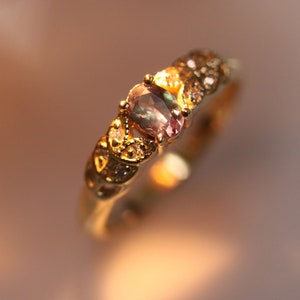 May include: A gold ring with a large oval-shaped gemstone set in the center. The gemstone is a light green and purple color with a shimmery finish. The ring is decorated with smaller clear gemstones around the center stone.