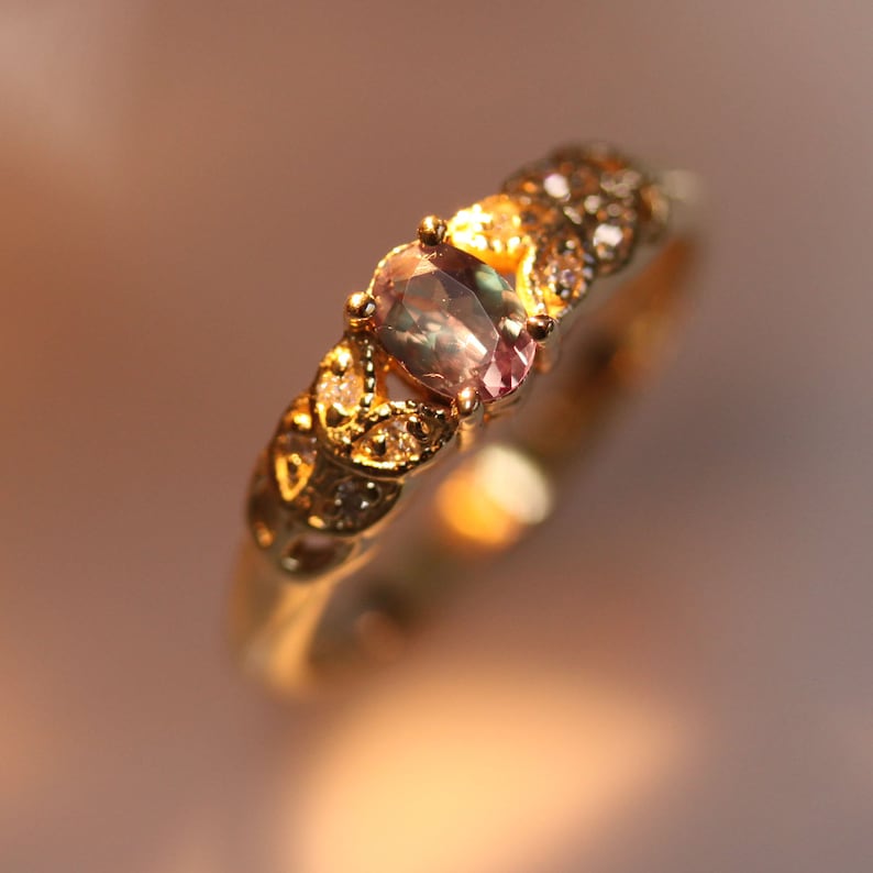 May include: A gold ring with a large oval-shaped gemstone set in the center. The gemstone is a light green and purple color with a shimmery finish. The ring is decorated with smaller clear gemstones around the center stone.