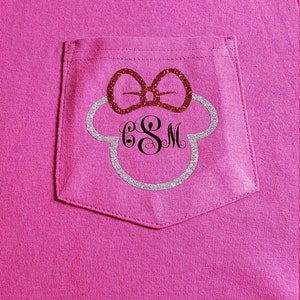 May include: Pink t-shirt with a pocket featuring a silver glitter Mickey Mouse head with a red glitter bow and the monogram "ESM" in black.