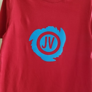 May include: Red t-shirt with a blue circle containing the letters "JV" in white. The circle is surrounded by a blue brushstroke design.