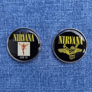 Puede incluir: Dos pines circulares de metal con fondo negro y bordes plateados. El pin izquierdo presenta el texto IN UTERO NIRVANA LIVE '94 y una figura alada. El pin derecho muestra el texto NIRVANA sobre un gráfico amarillo de una figura con alas.