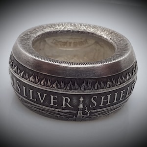 Puede incluir: Un anillo de plata con un diseño en relieve de llamas y las palabras "SILVER SHIELD".
