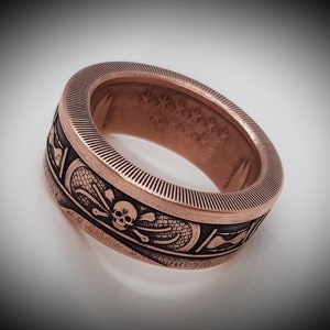 Könnte beinhalten: Ein kupferfarbener Ring mit einem Totenkopf- und Knochenkreuz-Design. Der Ring ist aus einer wiederverwendeten Münze gefertigt und hat eine strukturierte Oberfläche.