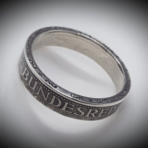 Può includere: Un anello d'argento realizzato con una moneta tedesca. L'anello presenta l'iscrizione "BUNDESREP" sul bordo esterno.