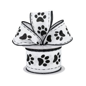 May include: White ribbon with black paw prints. The ribbon is decorated with a black stitched edge. The ribbon is arranged in a bow shape, with the spool visible below. The paw prints are of varying sizes.