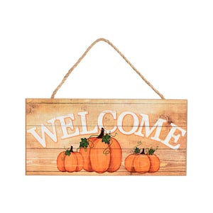 May include: A rectangular wooden sign with a light brown wood grain finish featuring the white text WELCOME across the center. Below the text are three orange pumpkins with green stems and leaves. A brown twine rope is attached for hanging.