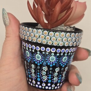 May include: A small, black painted flower pot with a dot art design in shades of blue, purple, and white. The rim is decorated with gold and white dots. The pot contains a faux succulent with reddish-brown leaves.
