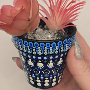 May include: A small, hand-painted black ceramic pot with a blue and white dot design, holding two faux succulents. One succulent is a dusty rose colour, and the other is a spiky, coral-coloured plant. The pot is filled with clear, decorative stones.