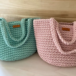 Op de afbeelding: Twee gebreide boodschappentassen, een in lichtgroen en de andere in lichtroze. Beide tassen hebben een bruin lederen label met de tekst "Handmade with Love" erop.