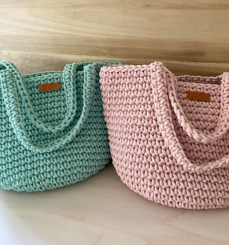 Op de afbeelding: Twee gebreide boodschappentassen, een in lichtgroen en de andere in lichtroze. Beide tassen hebben een bruin lederen label met de tekst "Handmade with Love" erop.