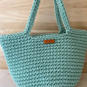 Op de afbeelding: Een lichtblauwe gehaakte tote bag met een bruin leren label waarop "Knit with Love" staat.