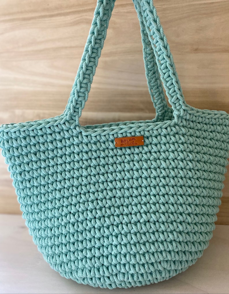 Op de afbeelding: Een lichtblauwe gehaakte tote bag met een bruin leren label waarop "Knit with Love" staat.