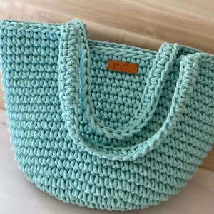 Op de afbeelding: Een lichtblauwe gehaakte tote bag met een bruin leren label waarop "Happy Chic" staat.