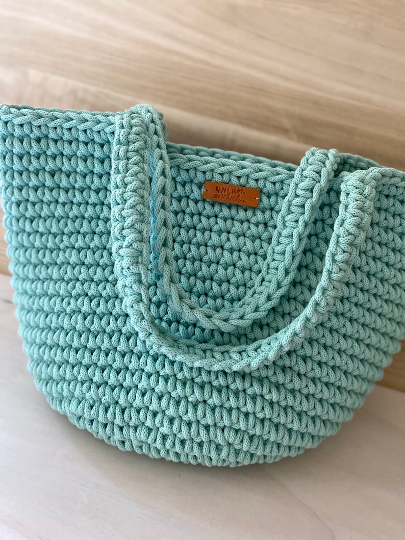 Op de afbeelding: Een lichtblauwe gehaakte tote bag met een bruin leren label waarop "Happy Chic" staat.
