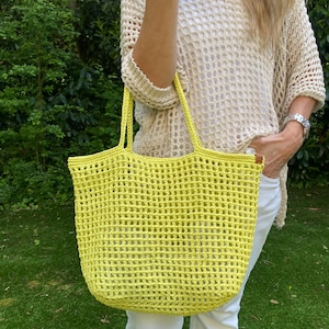 Peut inclure: Un sac fourre-tout crocheté jaune vif avec un motif de tissage ouvert rectangulaire. Le sac a deux anses et est tenu par une personne portant un pull crème et un pantalon blanc. L'arrière-plan est de l'herbe verte et du feuillage.