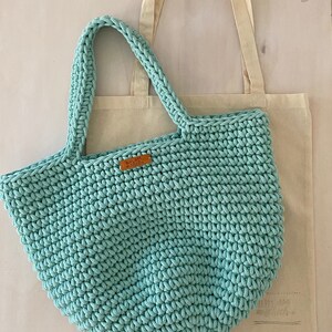 Op de afbeelding: Een lichtblauwe gehaakte tote bag met een bruin leren label waarop "Handmade with Love" staat. De tas ligt op een witte canvas tote bag.