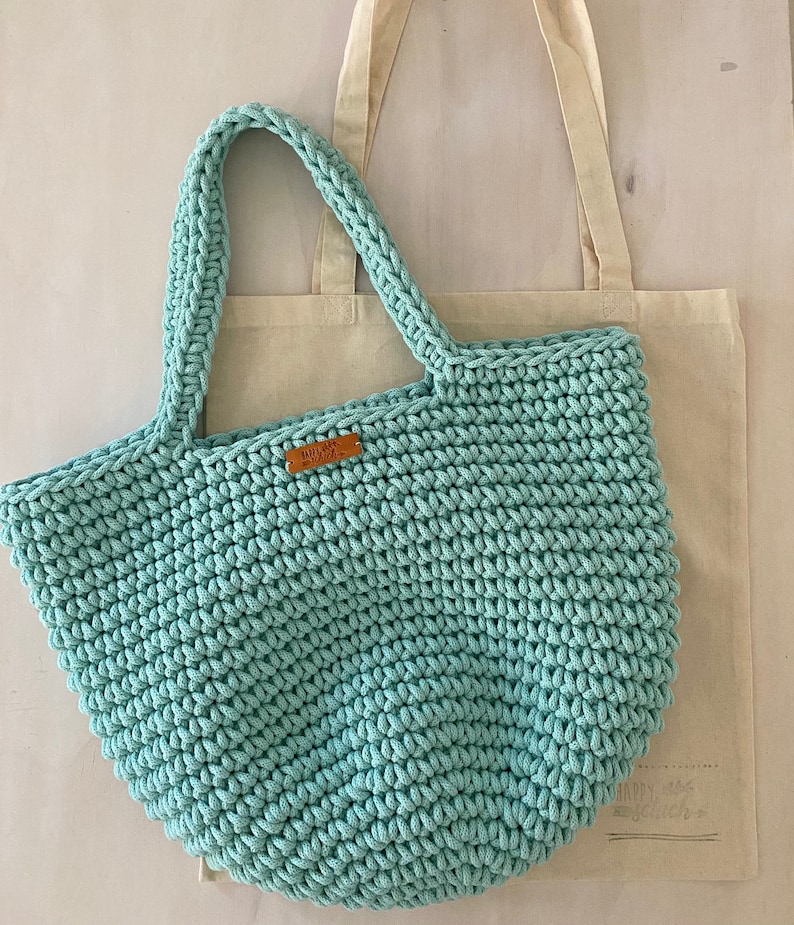 Op de afbeelding: Een lichtblauwe gehaakte tote bag met een bruin leren label waarop "Handmade with Love" staat. De tas ligt op een witte canvas tote bag.