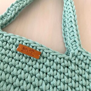 Op de afbeelding: Een lichtgroene gehaakte tote bag met een bruin leren label waarop "Happy Chic" staat.
