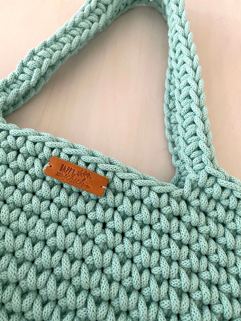 Op de afbeelding: Een lichtgroene gehaakte tote bag met een bruin leren label waarop "Happy Chic" staat.