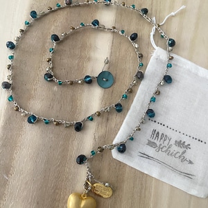 Puede incluir: Un collar hecho a mano con una cadena plateada, adornado con cuentas azules, turquesas y doradas. Un colgante de corazón dorado y una pequeña etiqueta cuelgan de la cadena. Un botón turquesa y una pequeña bolsa blanca con el texto "HAPPY schich" también son visibles.