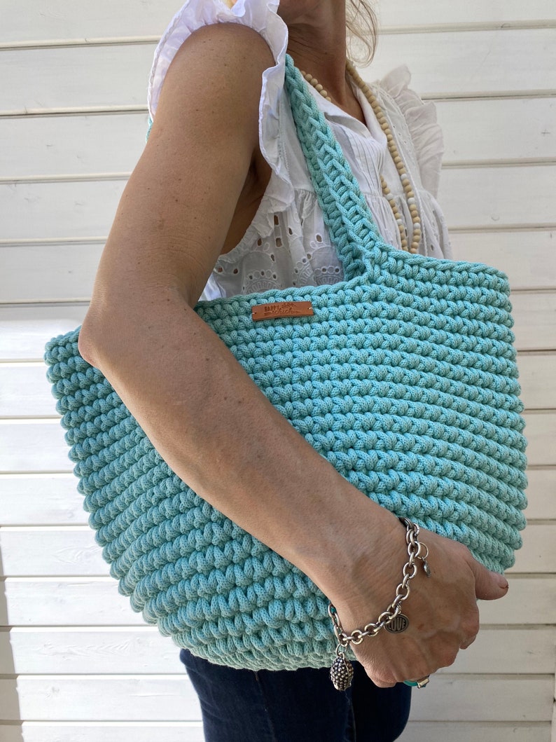 Op de afbeelding: Een lichtblauwe gehaakte tote bag met een leren label waarop "Happy Hour" staat. De tas heeft een brede, platte bodem en twee lange hengsels.