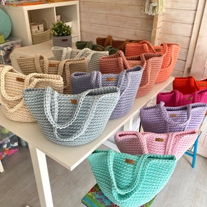 Op de afbeelding: Een verzameling gehaakte tassen in verschillende kleuren, waaronder lichtblauw, lavendel, roze en groen. De tassen zijn gerangschikt op een witte tafel en een houten kruk.
