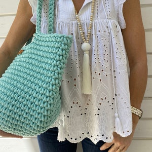 Op de afbeelding: Een lichtblauwe gehaakte tote bag met een witte kwast. De tas wordt gedragen door iemand die een wit kanttopje en een blauwe jeans draagt.