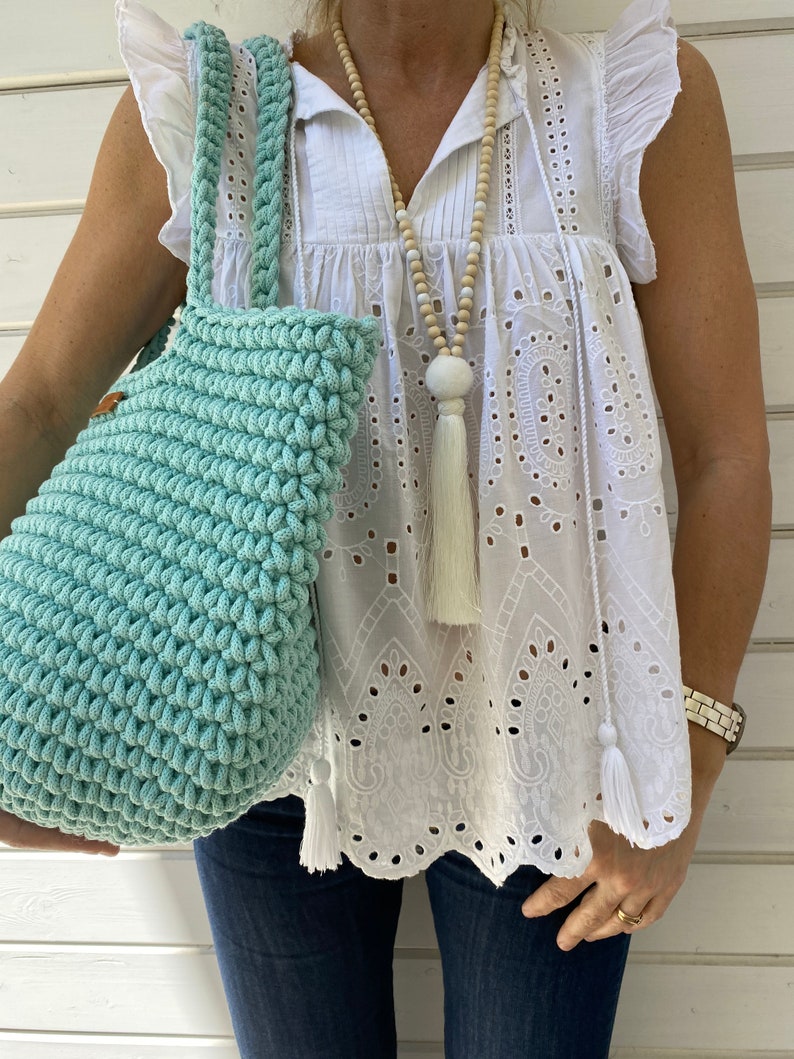 Op de afbeelding: Een lichtblauwe gehaakte tote bag met een witte kwast. De tas wordt gedragen door iemand die een wit kanttopje en een blauwe jeans draagt.