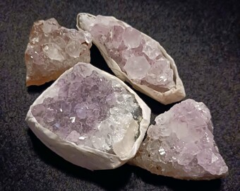 May include: Five amethyst crystal clusters in shades of purple and white. The crystals have a natural, rough texture and are set against a dark background. The crystals vary in size and shape, showcasing their unique formations.